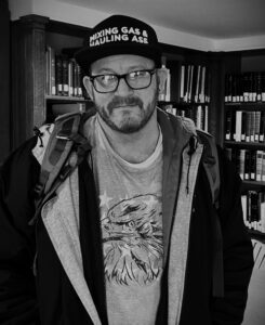 Black and white photograph of David Thompson wearing a graphic tee, a baseball hat, and dark-rimmed glasses.